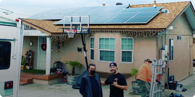 Two men pose in front of a house equipped with solar panels, highlighting sustainable living and energy efficiency.